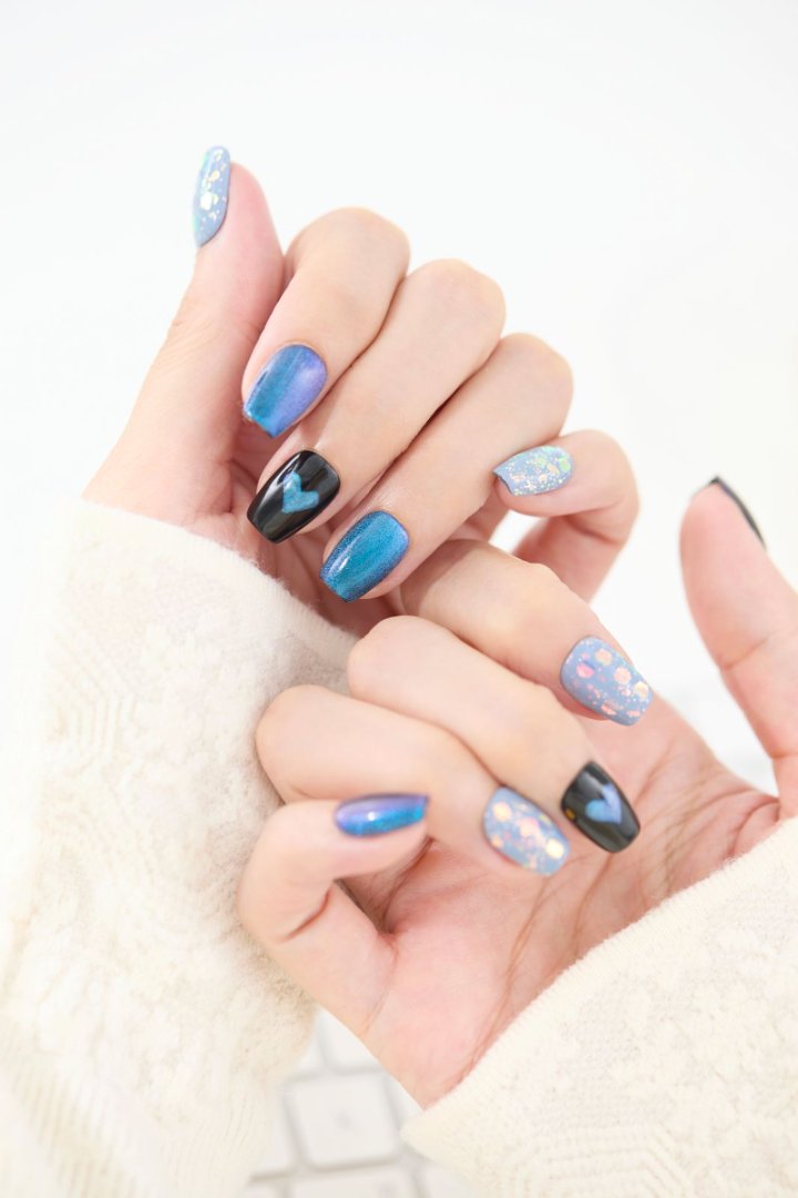 「ブルーマグネット×ブラックハートのジェルネイルチップを指先に着けた手元アップ。オーロラフレークのブルーネイルが光に輝くリユーザブルネイルデザイン」