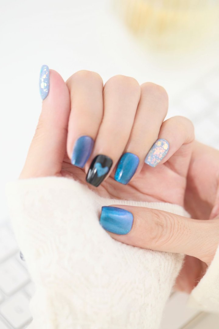 「ブルーマグネット×ブラックハートのジェルネイルチップを指先に着けた手元アップ。オーロラフレークのブルーネイルが光に輝くリユーザブルネイルデザイン」